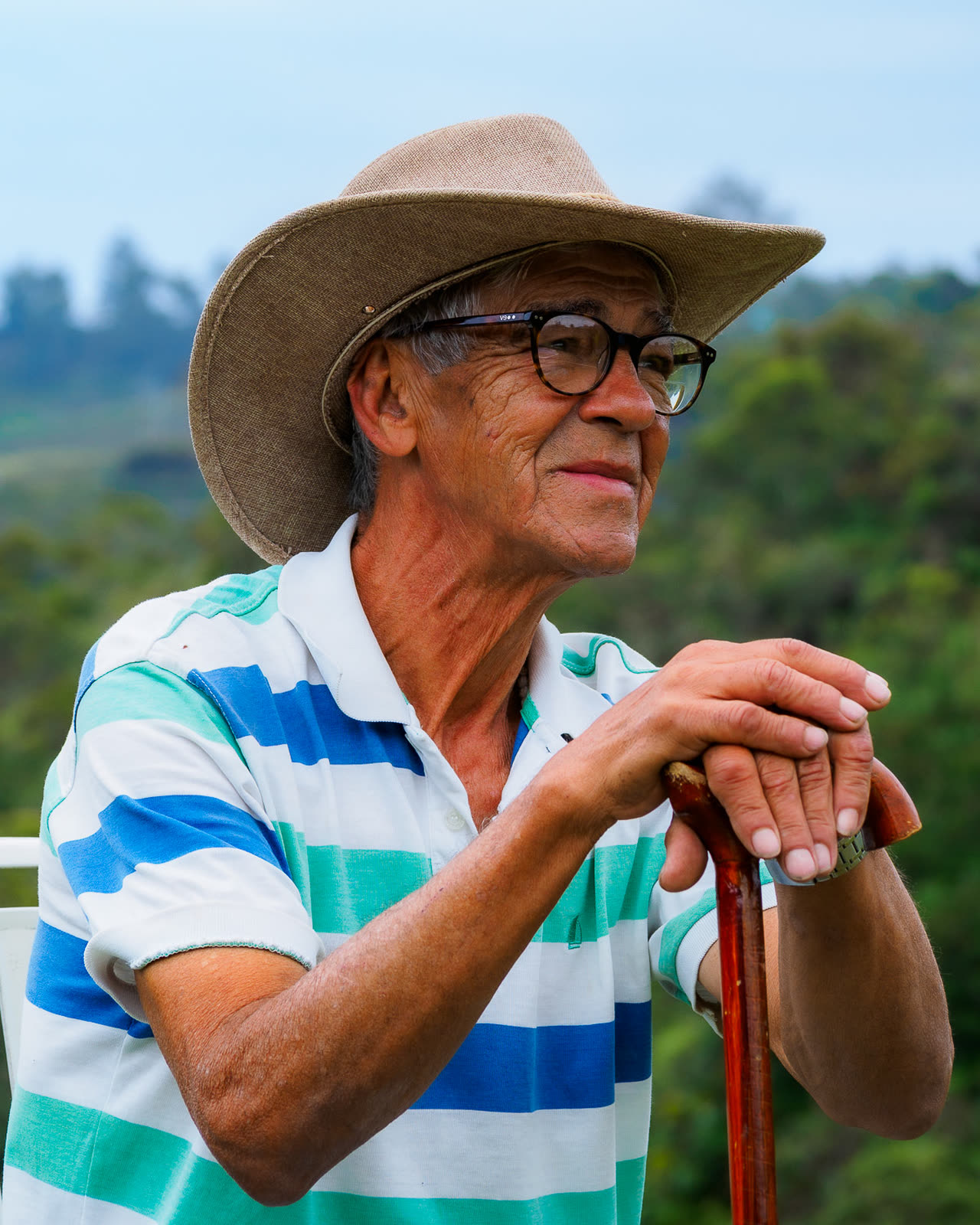 Retrato de un hombre mayor con sombrero apoyado en su bastón, en un paisaje verde