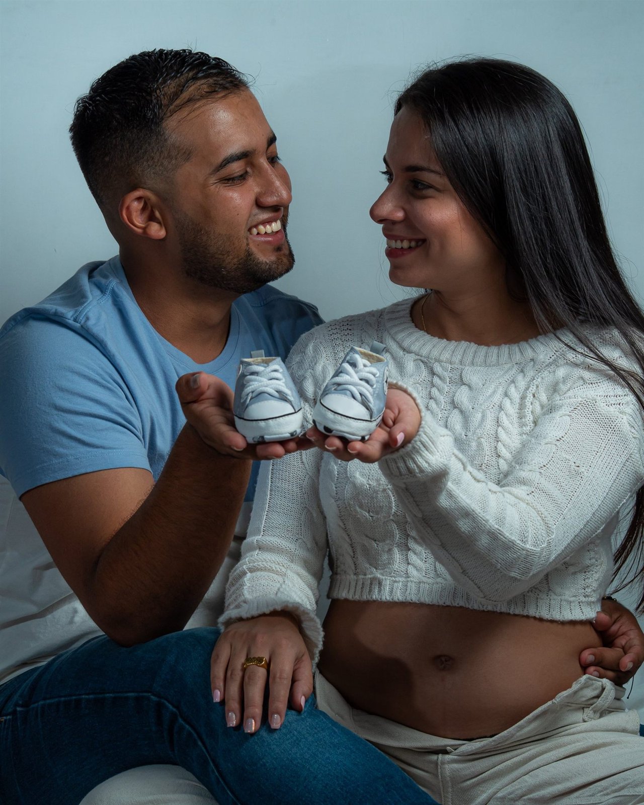 Pareja embarazada sonriéndose mientras sostienen unos zapatitos de bebé