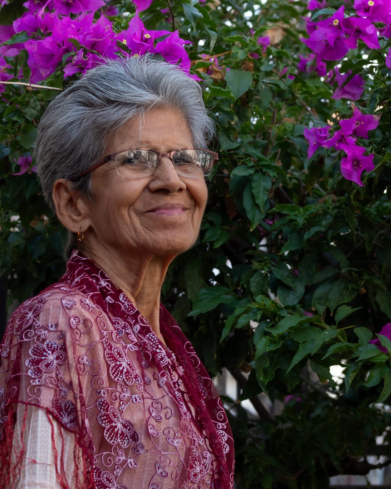 Mujer mayor de cabello gris y gafas sonriendo ante una buganvilla en flor