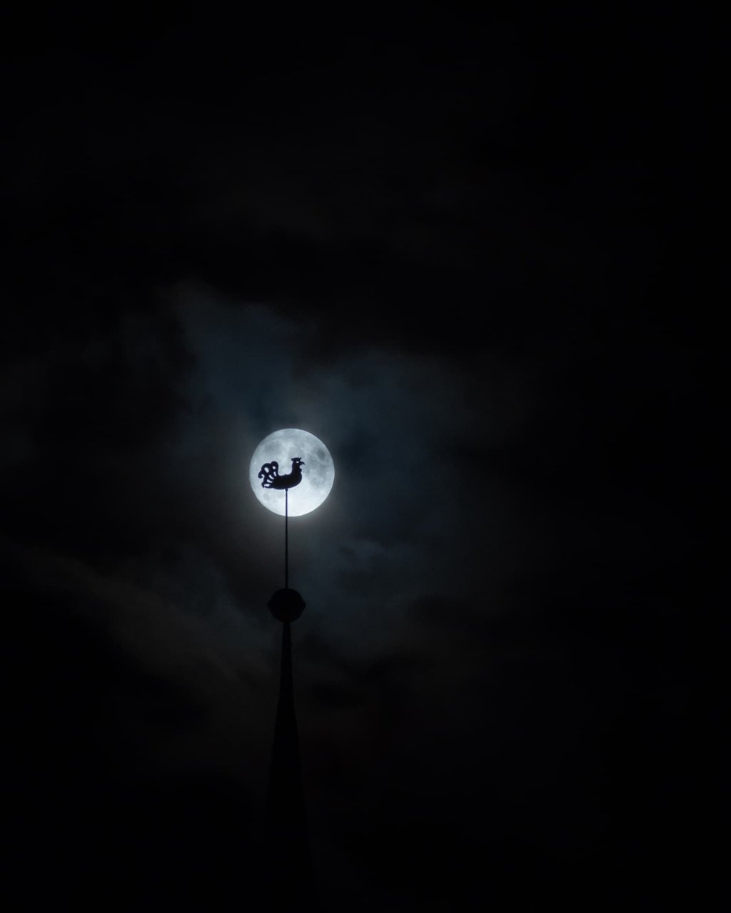 Luna llena tras una veleta con forma de gallo en lo alto de una aguja, en blanco y negro