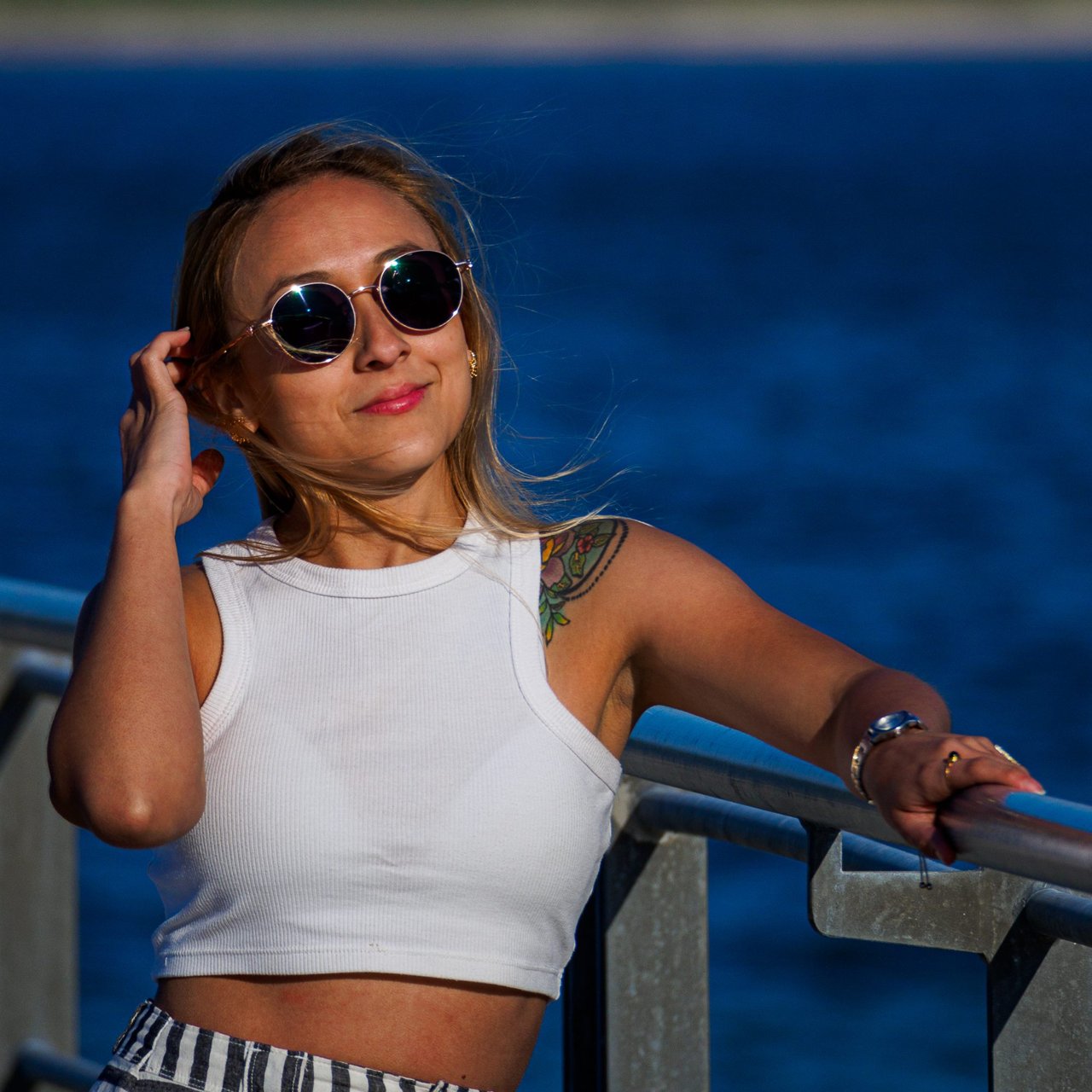 Mujer con gafas de sol sonriendo apoyada en una barandilla frente al mar azul