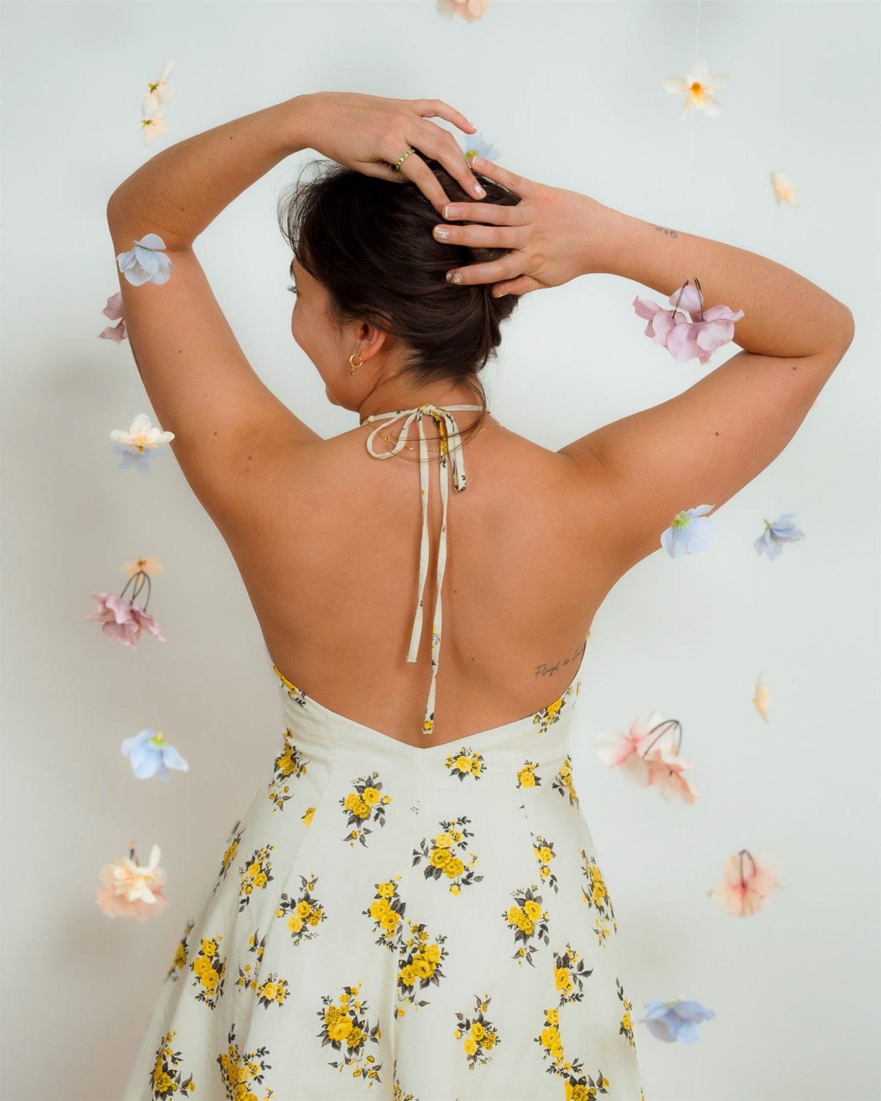 Mujer de espaldas con vestido de flores y manos en la cabeza ante una pared con flores de papel