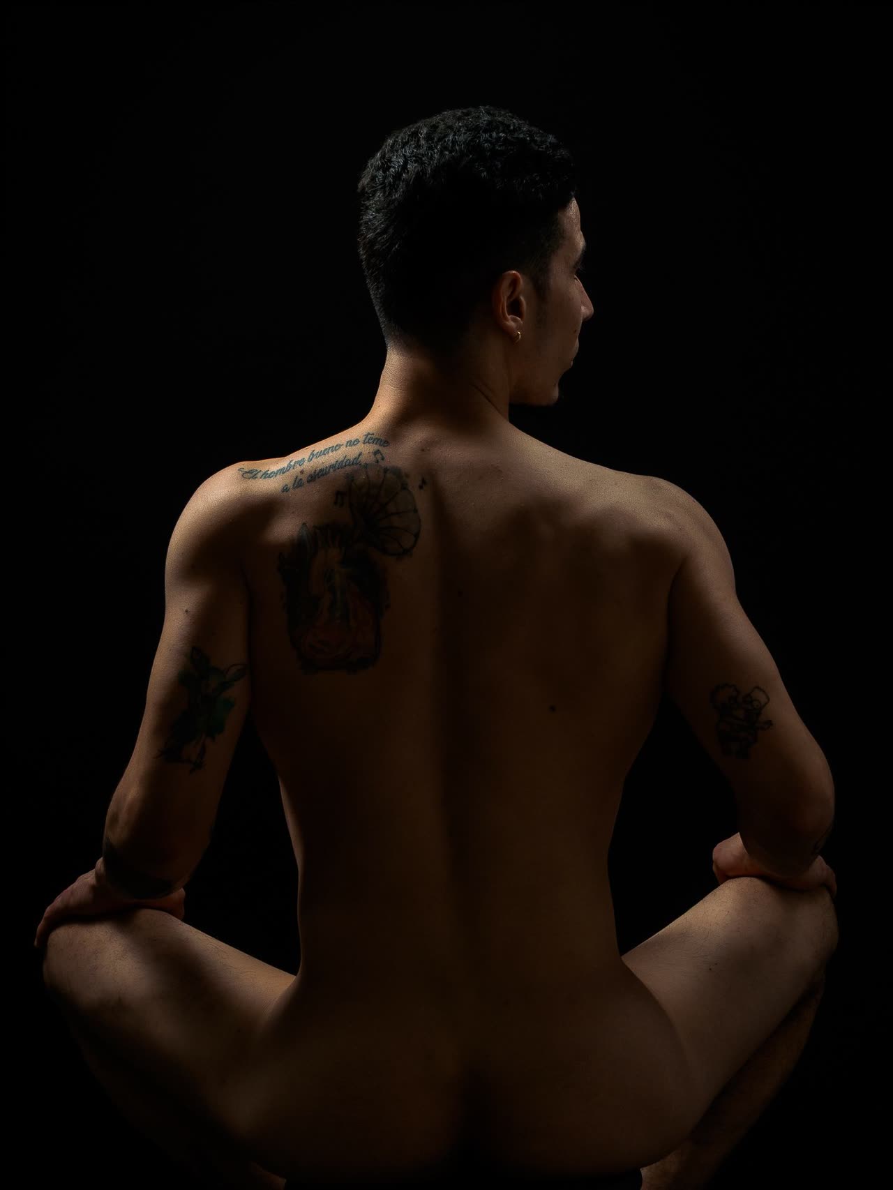 Hombre de espaldas con tatuajes, sentado en cuclillas sobre fondo negro