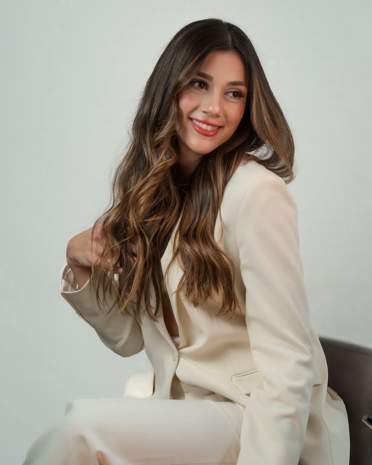 Mujer sonriente de cabello largo con traje claro, sentada ante un fondo gris suave