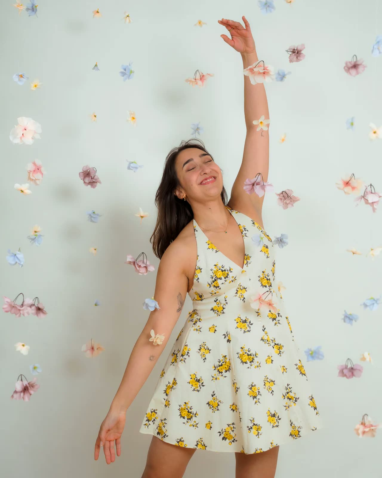 Mujer con vestido de flores levantando un brazo ante una pared de flores de papel